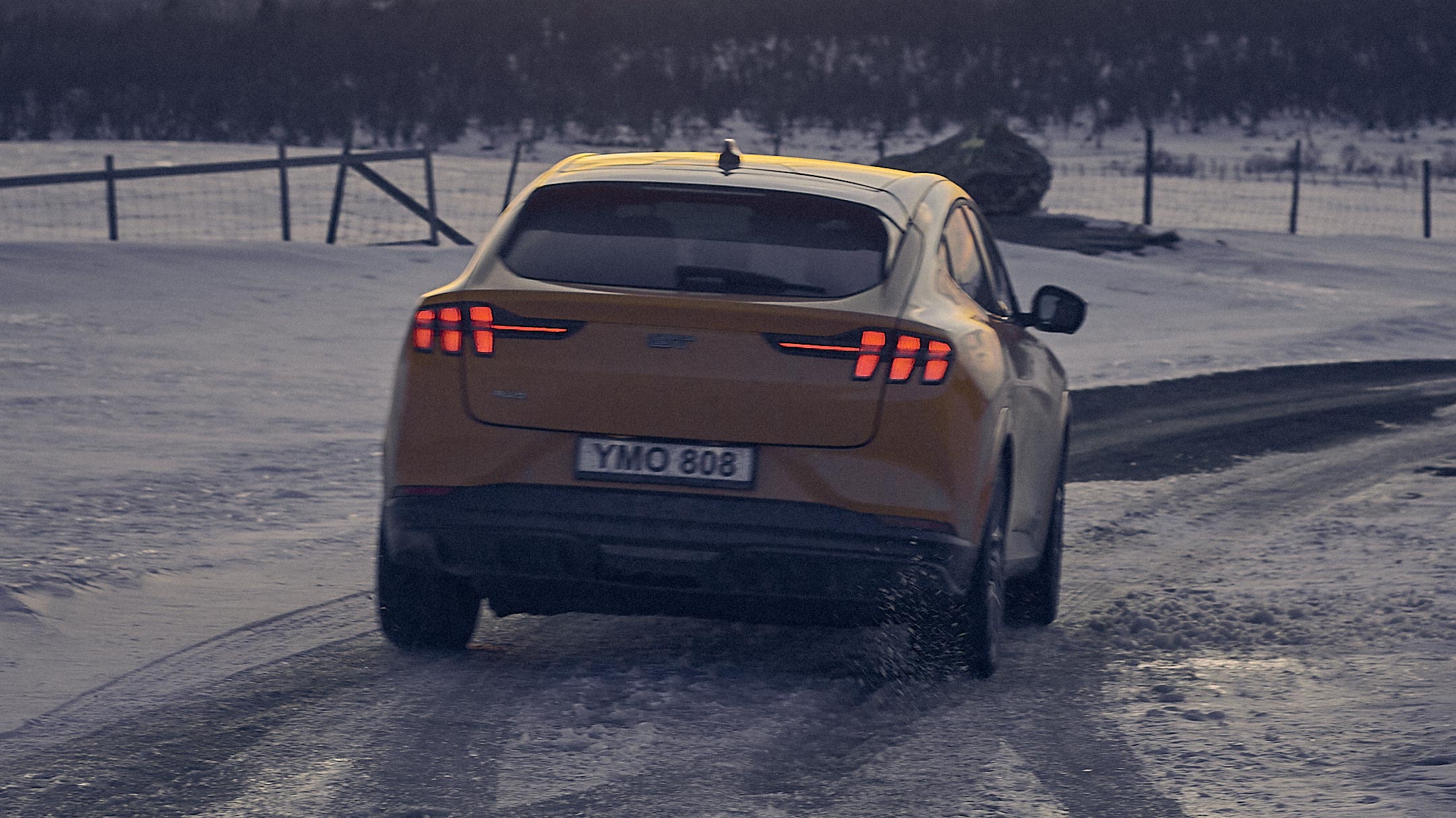 Ford Mustang Mach-E in Orange. Heckansicht, fahrend auf einer mit Schnee bedeckten Straße.
