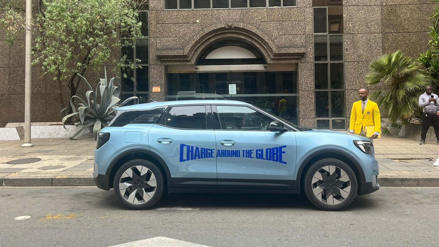 Vollelektrischer Ford Explorer in Blau. Seitenansicht mit „Charge Around The Globe“-Logo, parkt vor einem Gebäude.