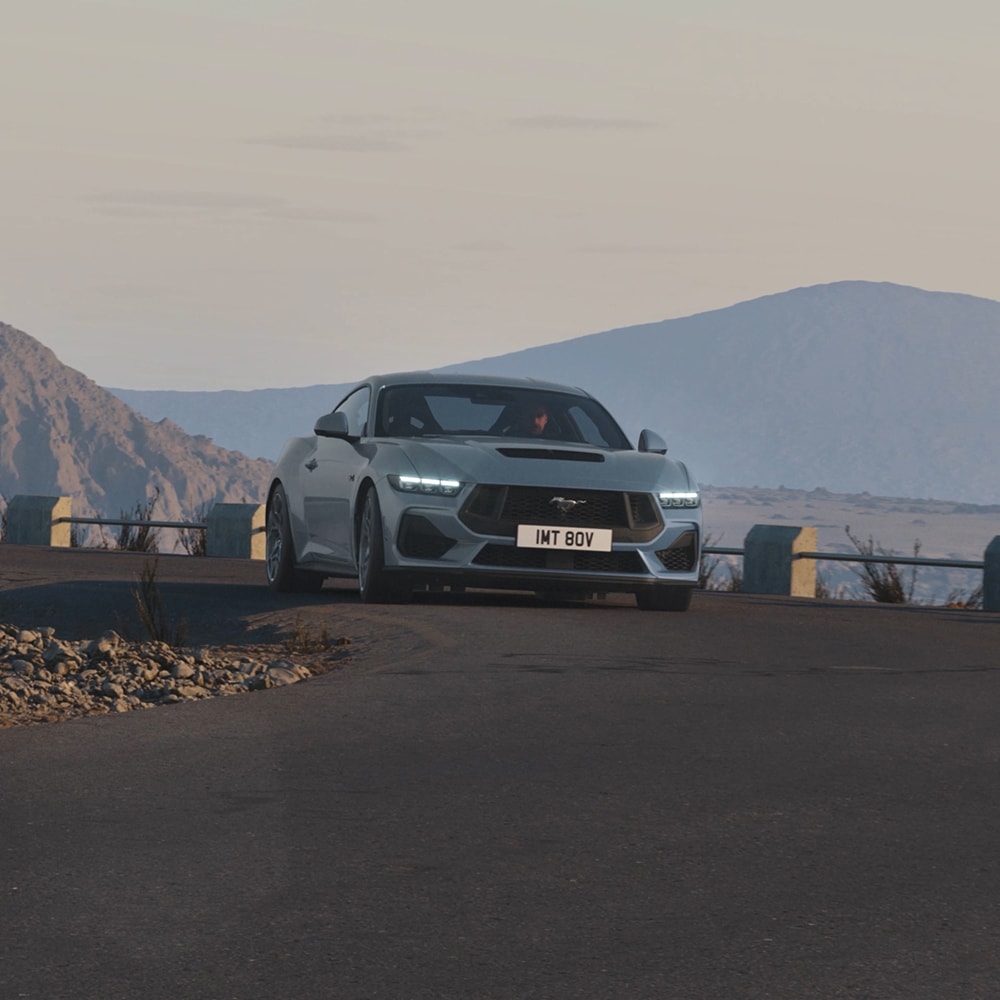 Ford Mustang in Blau. Frontansicht, fahrend auf einer Straße mit Bergen im Hintergrund.