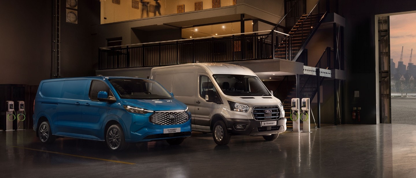 Ford E-Transit Custom in Blau und Ford E-Transit in Silber parken nebeneinander in einer Halle neben Ladesäulen.