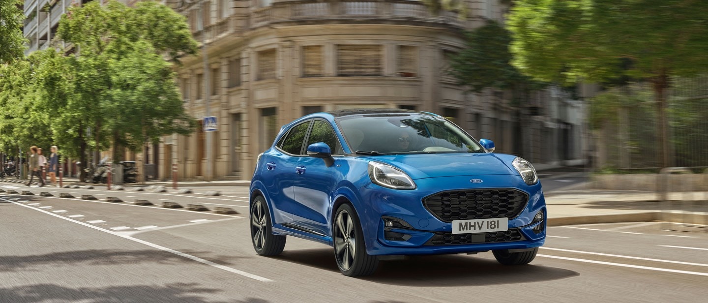 Ford Puma in Blau. Dreiviertelansicht, fahrend in Stadt über Kreuzung. 