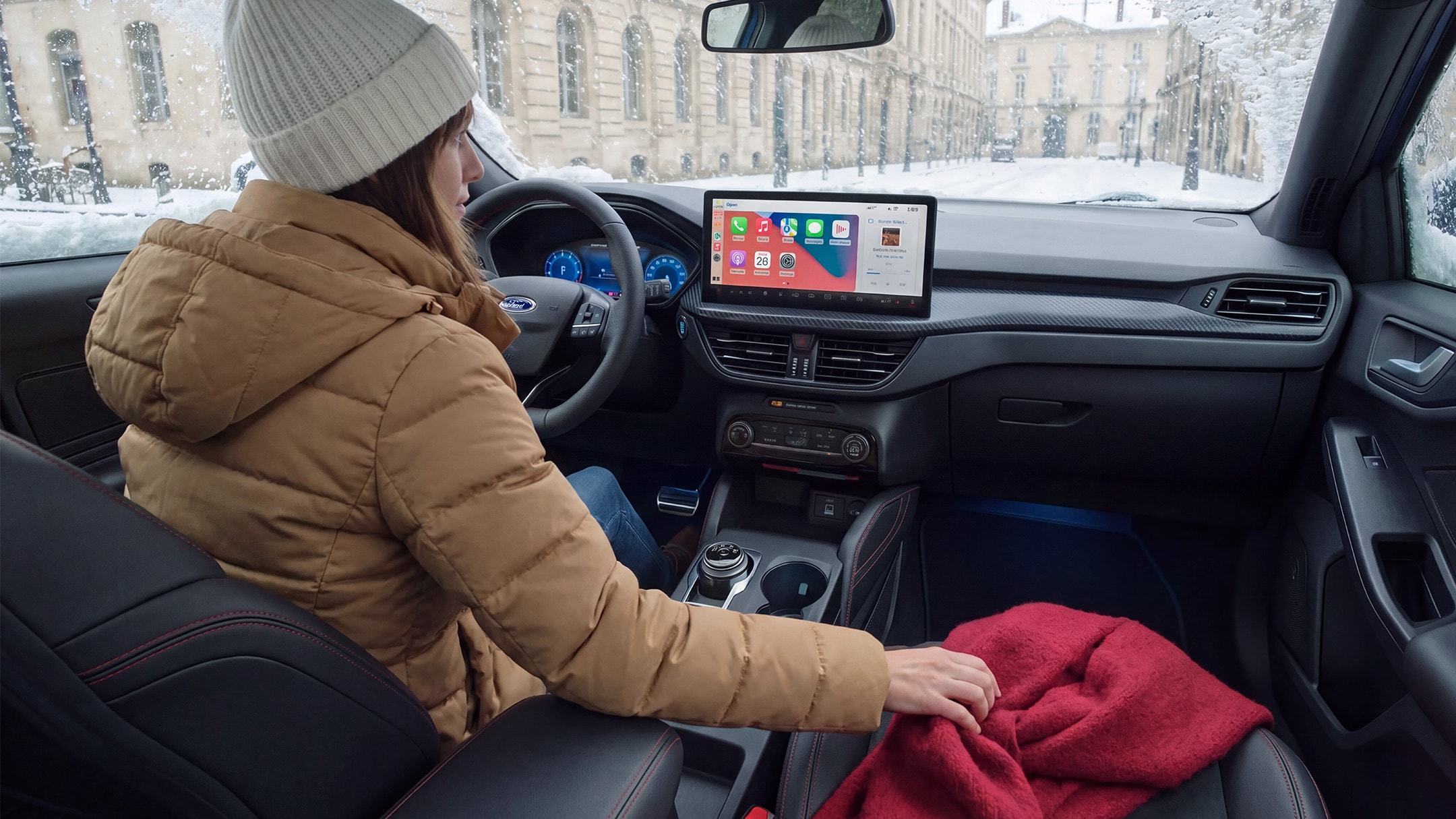 Eine Frau in Wintermantel und Haube sitzt an einem verschneiten Tag in einem Ford und greift nach einer roten Decke.