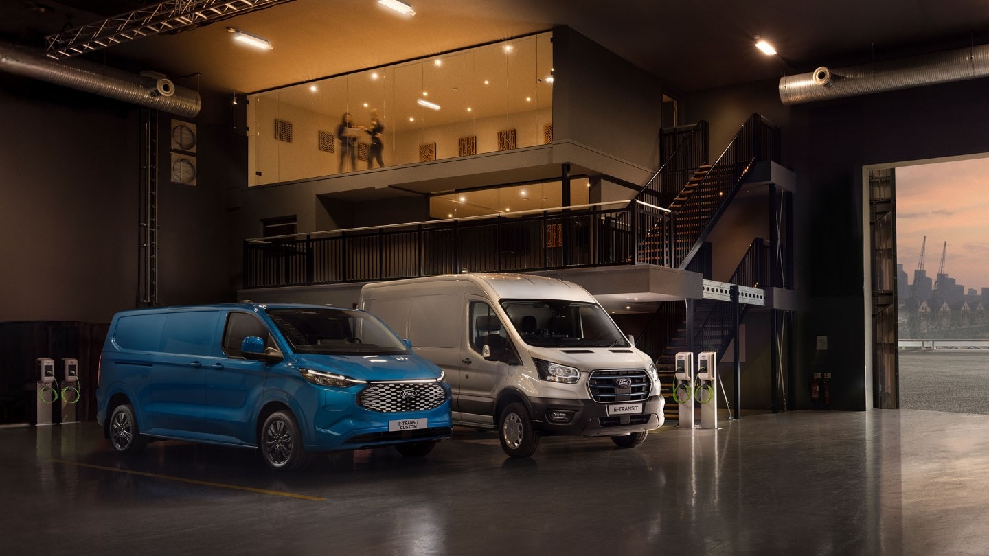 Ford E-Transit Custom in Blau und Ford E-Transit in Silber parken nebeneinander in einer Halle neben Ladesäulen. 