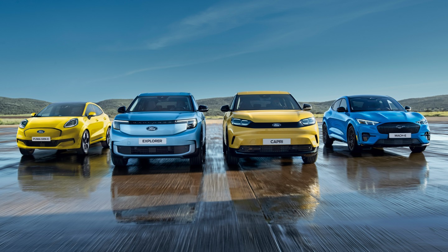 Puma Gen-E, Explorer, Capri und Mustang Mach-E stehen auf einer nassen Fahrbahn. Frontansicht der Fahrzeuge. 