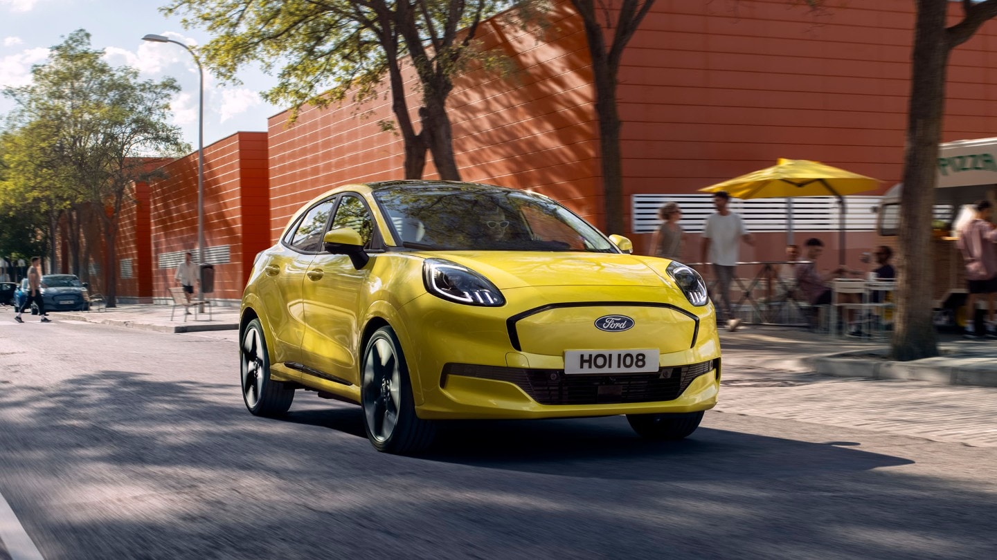 Ford Puma Gen-E in Gelb. Dreiviertelansicht, fährt auf einer Straße mit Bäumen und Personen im Hintergrund.
