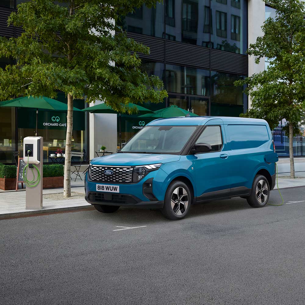 Ford E-Transit Courier in Blau. Dreiviertelansicht, parkt in einer Innenstadt und lädt an einer Ladestation.