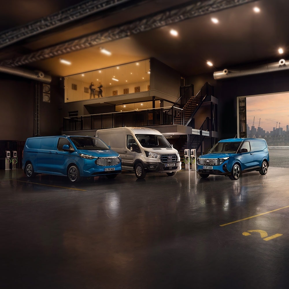 Ford E-Transit Custom in Blau und Ford E-Transit in Silber parken nebeneinander in einer Halle neben Ladesäulen.