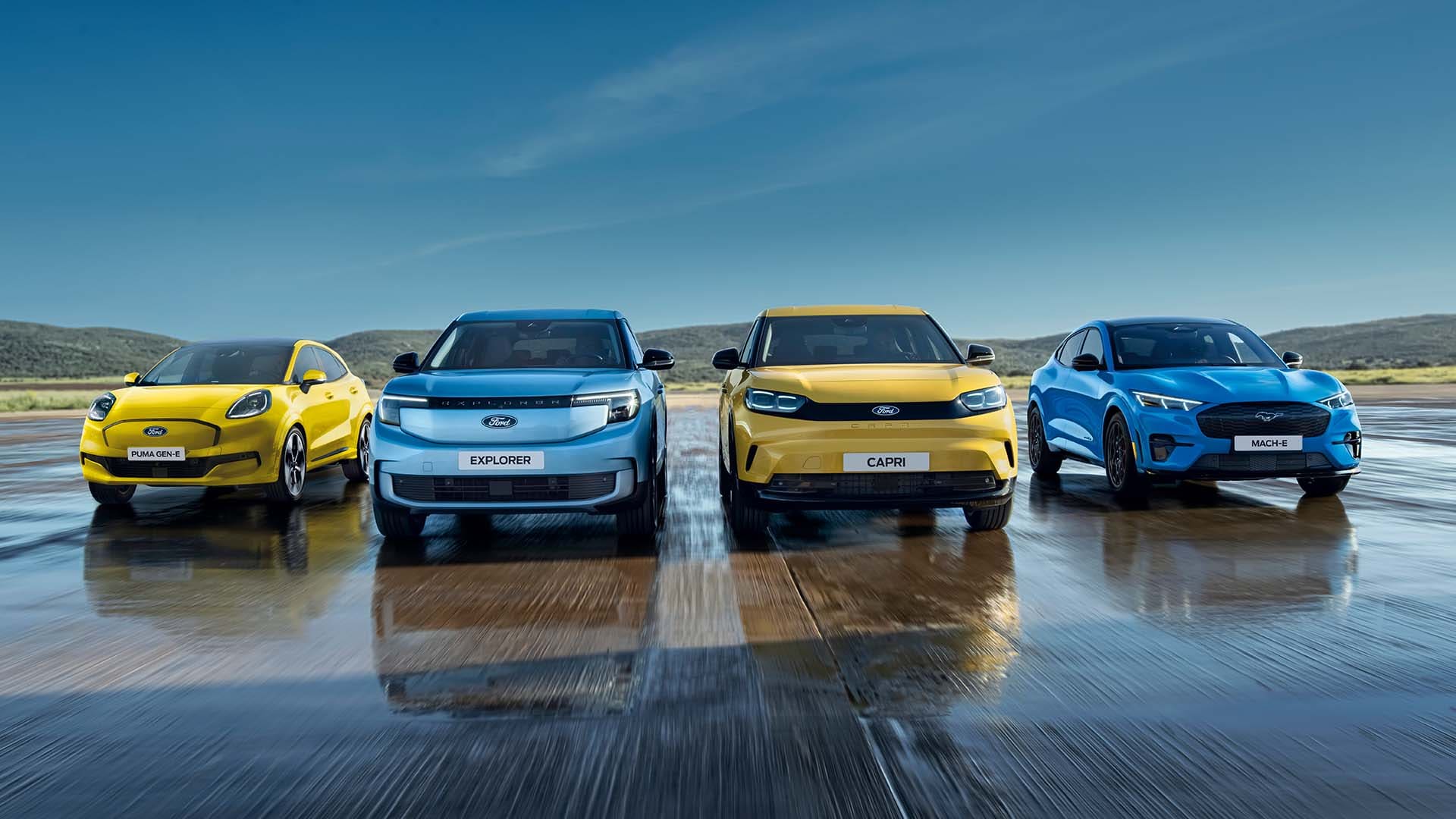 Puma Gen-E, Explorer, Capri und Mustang Mach-E stehen auf einer nassen Fahrbahn. Frontansicht der Fahrzeuge.