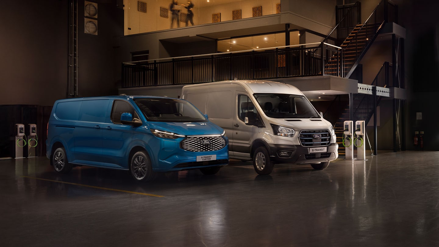 Ford E-Transit Custom in Blau und Ford E-Transit in Silber parken nebeneinander in einer Halle neben Ladesäulen.