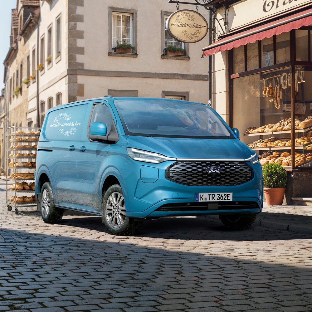 Ford E-Transit Custom in Blau. Dreiviertelansicht, parkt auf der Straße vor einer Bäckerei.
