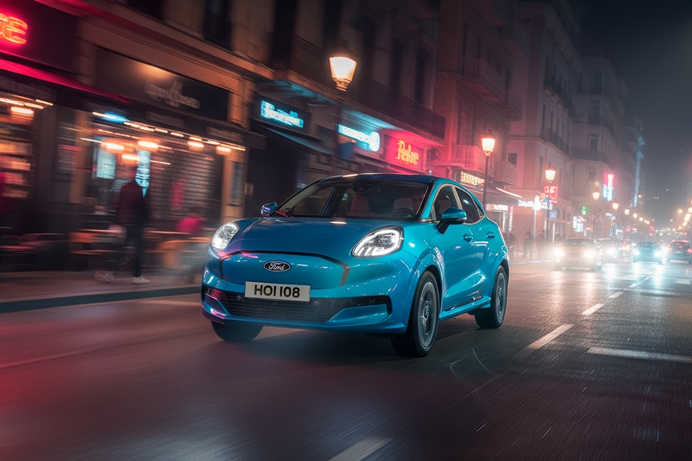Ford Puma in Blau. Dreiviertelansicht, fährt bei Nacht durch eine Stadt mit beleuchteten Geschäften. 