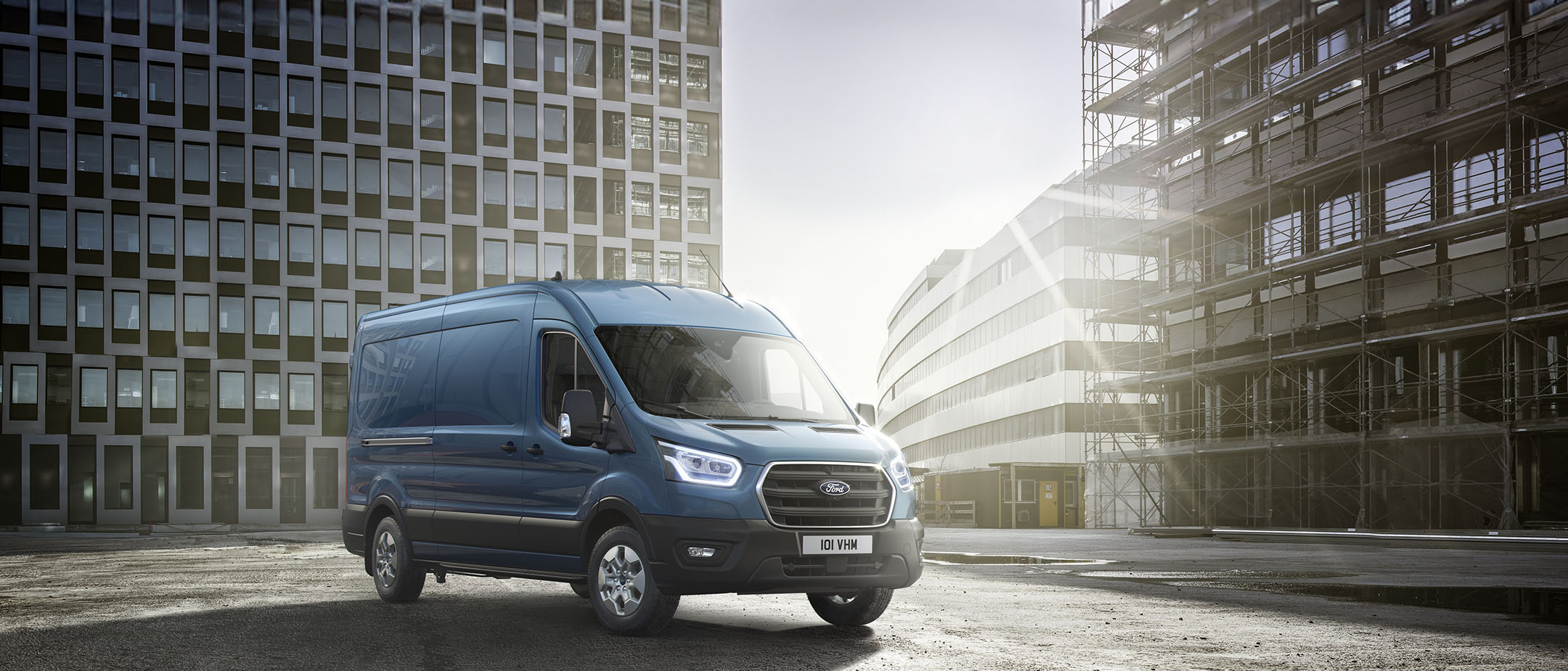 Ford Transit in Blau. Dreiviertelansicht, parkt vor einem modernen Gebäude mit einem Baugerüst.