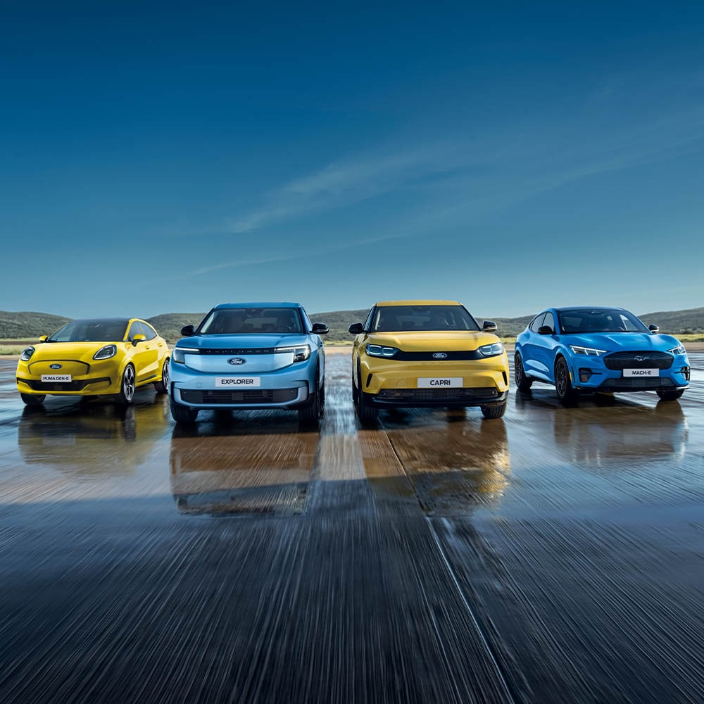 Puma Gen-E, Explorer, Capri und Mustang Mach-E stehen auf einer nassen Fahrbahn. Frontansicht der Fahrzeuge.