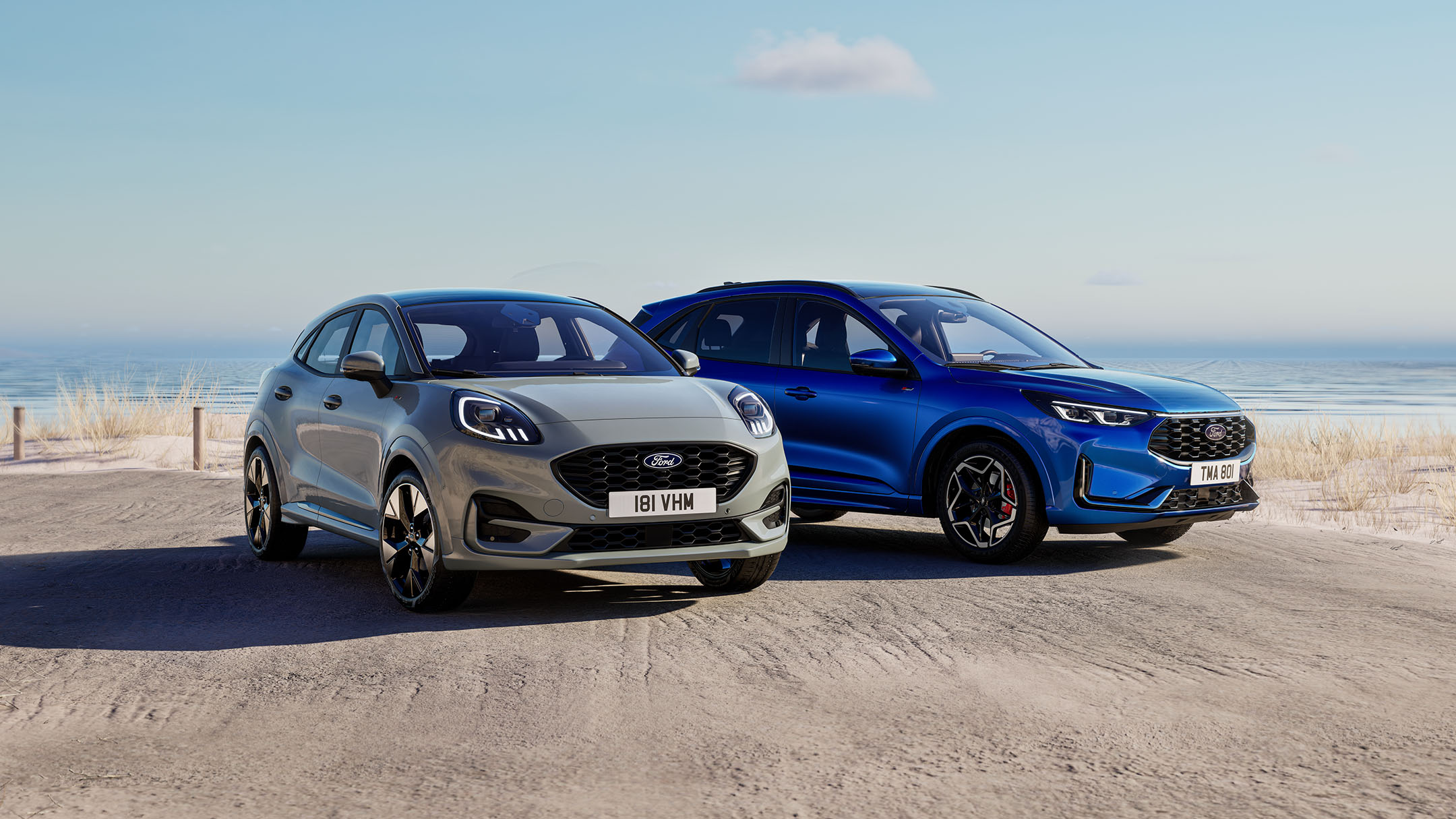 Ein grauer Ford Puma und blauer Ford Kuga parken nebeneinander auf einem Parkplatz neben dem Meer.