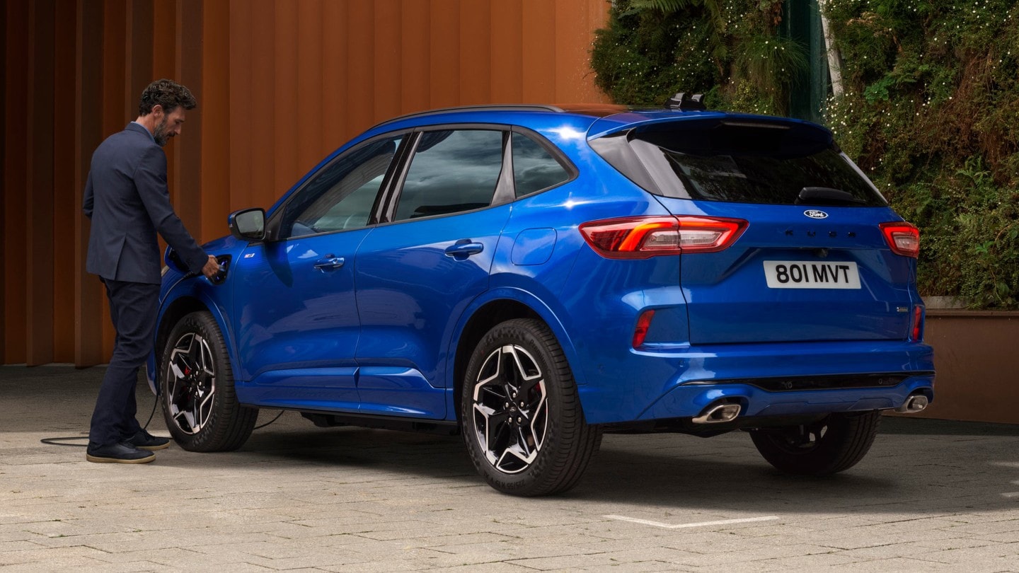 Ford Kuga in Blau. Heckansicht, Mann schließt Ladekabel an das Fahrzeug an.