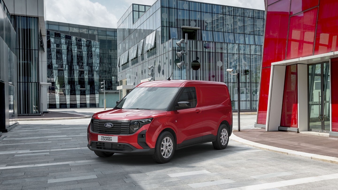 Ford Transit Courier in Rot. Dreiviertelansicht, geparkt vor einem Gebäude mit Glasfront.