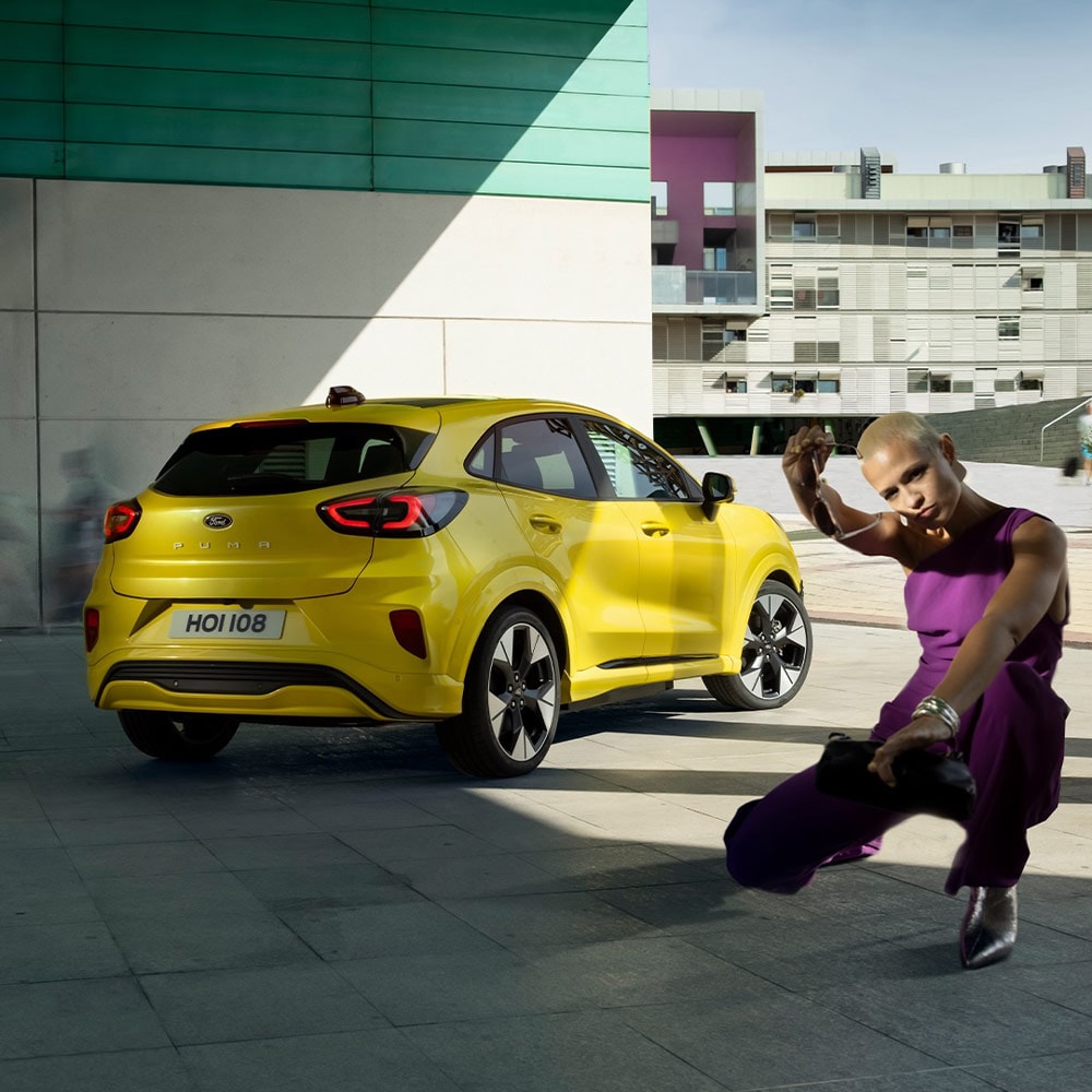 Ford Puma Gen-E in Gelb. Heckansicht, parkt in einem Wohngebiet. Eine Person sitzt in der Hocke vor dem Elektro-SUV.
