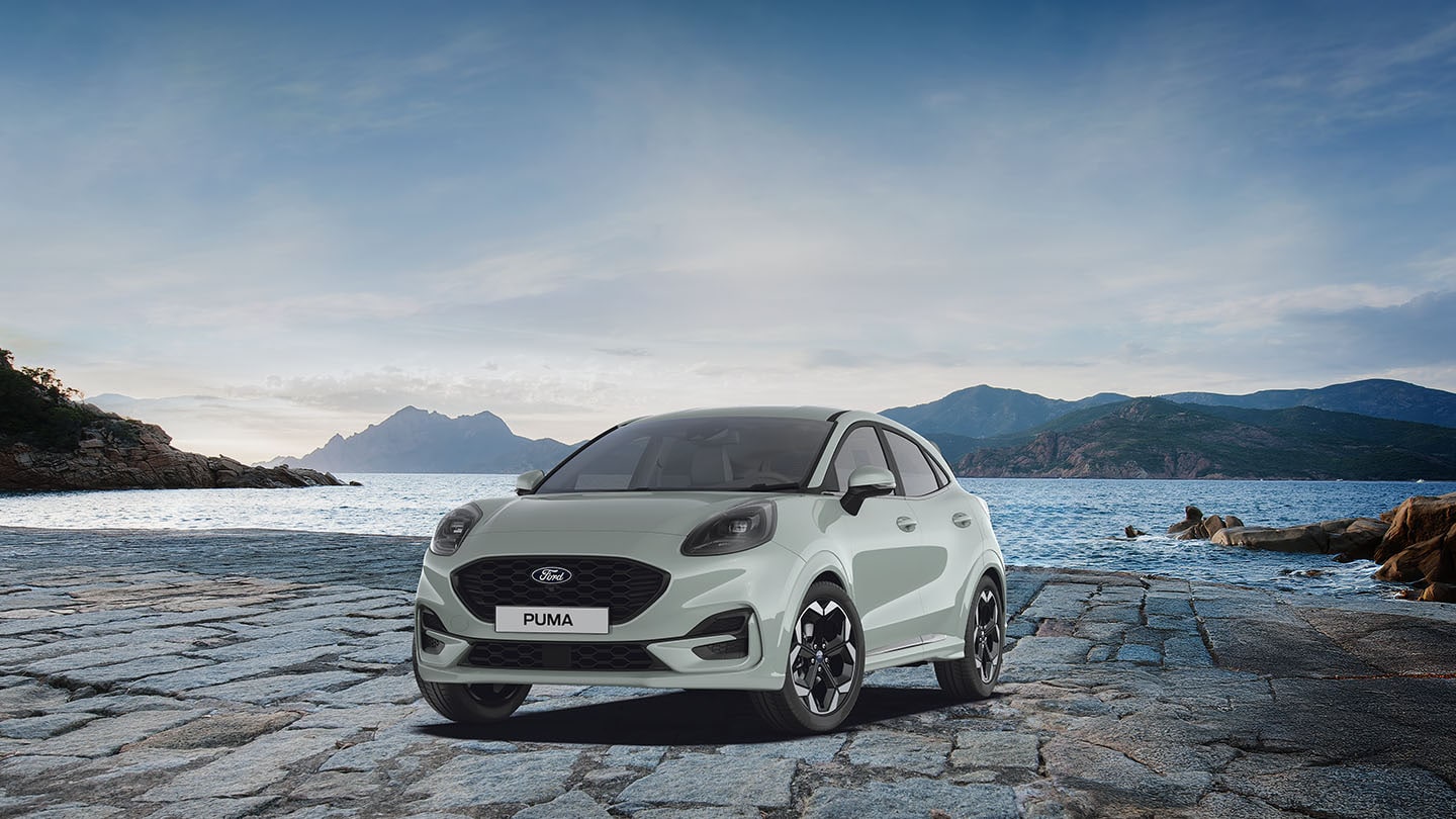 Ford Puma in Grau. Dreiviertansicht, parkt vor einem See mit Berglandschaft.