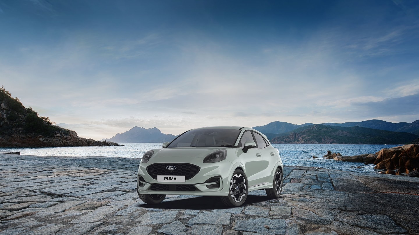 Ford Puma in Grau. Dreiviertansicht, parkt vor einem See mit Berglandschaft.