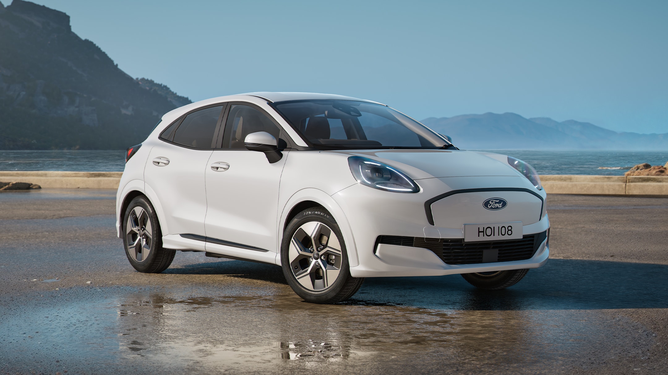 Ford Puma in Weiß. Dreiviertelansicht, parkt auf einer nassen Fahrbahn mit Hügellandschaft im Hintergrund. 