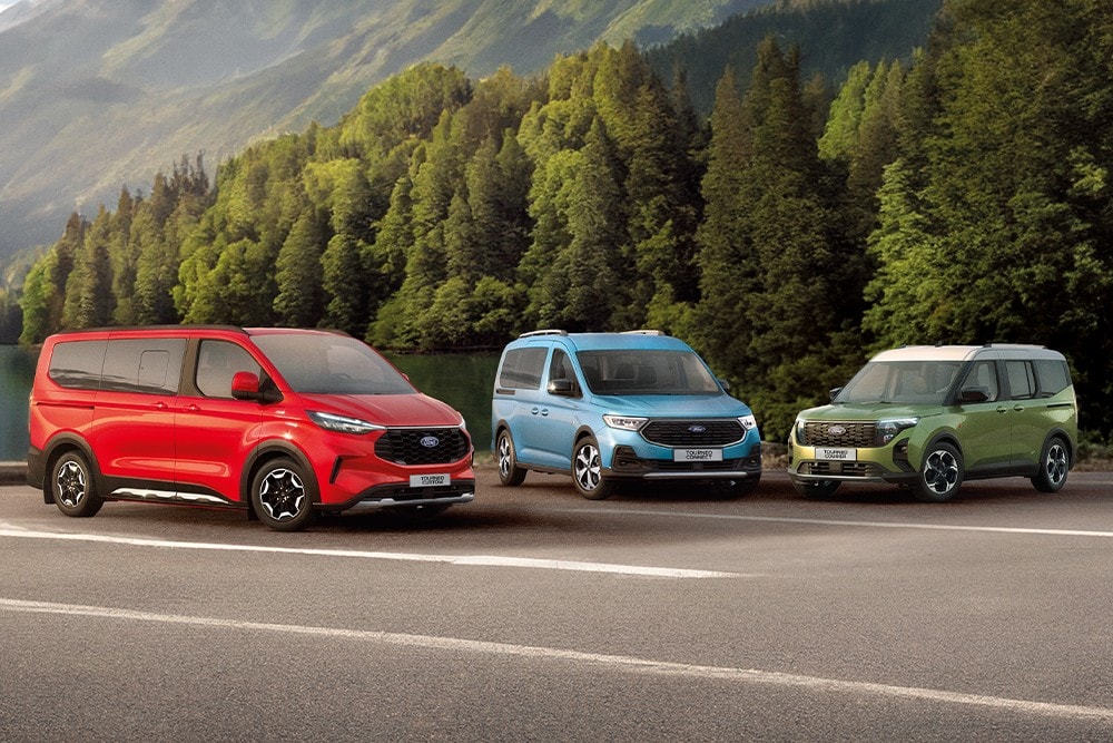 Ein roter Ford Tourneo Custom, ein blauer Tourneo Connect und ein grüner Tourneo Courier stehen nebeneinander auf einem Parkplatz, im Hintergrund ein grüner Wald.