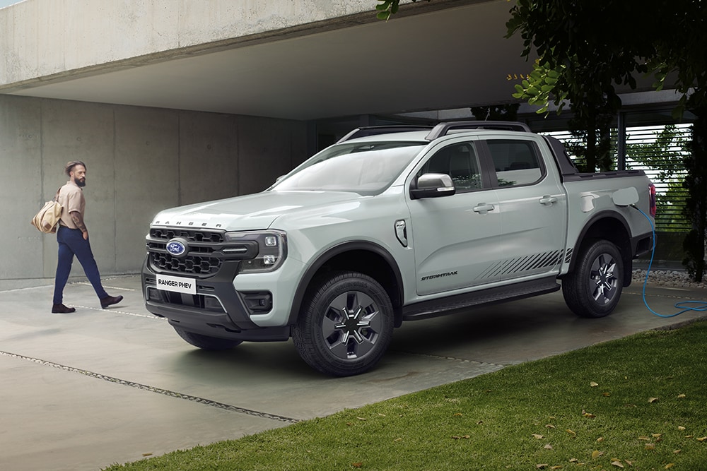 Ford Ranger Plug-in-Hybrid in Grau. Dreiviertelansicht, parkend vor einem Wohnhaus. Mann geht an Fahrzeug vorbei.