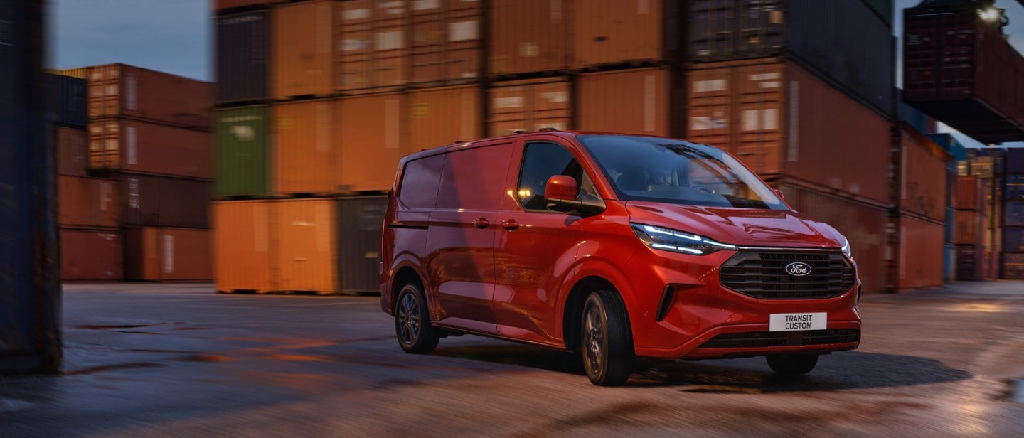 Ford Transit Custom in Rot. Dreiviertelansicht, fahrend auf einer Straße mit Containern im Hintergrund.