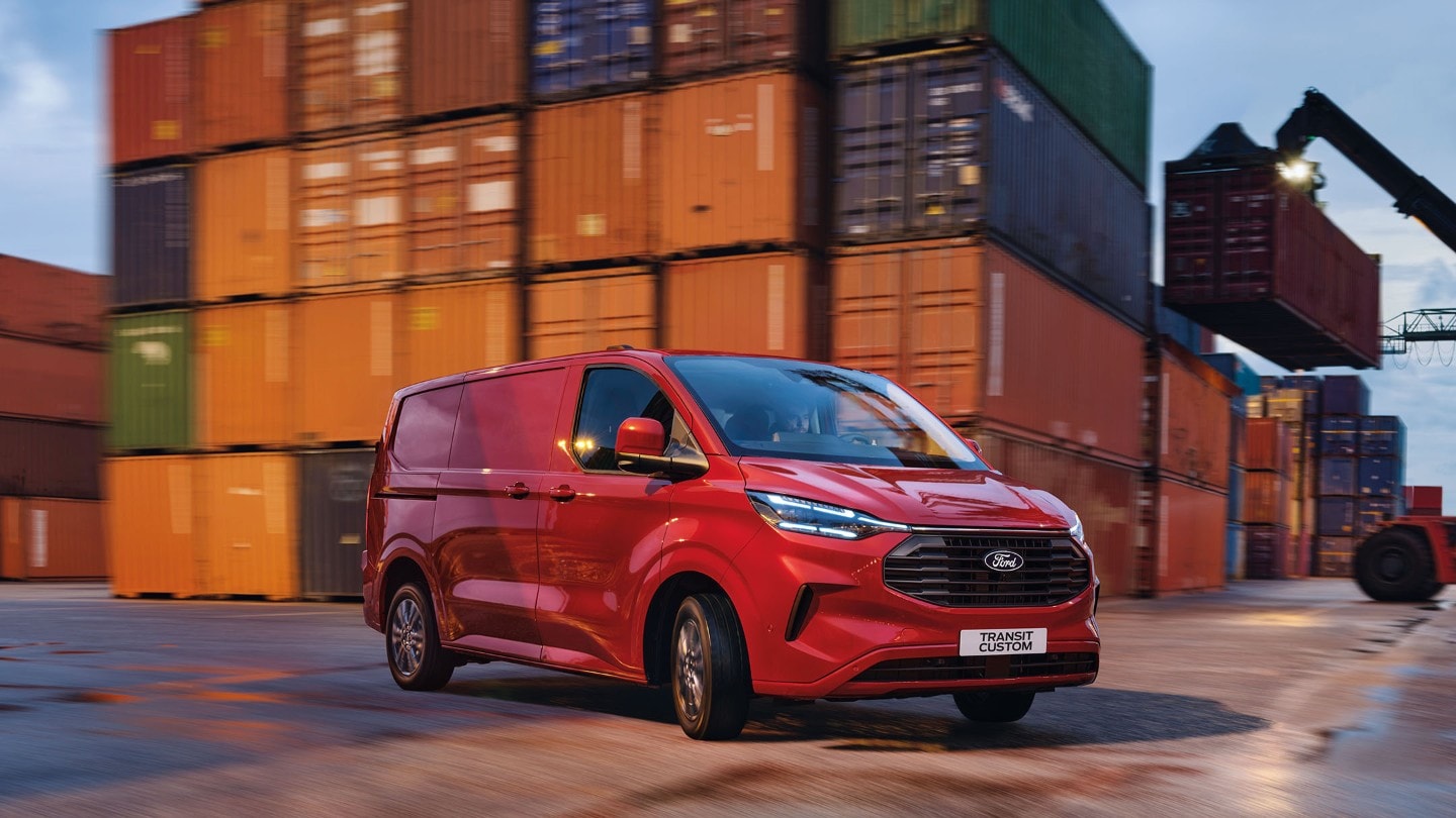 Ford Transit Custom in Rot. Dreiviertelansicht, fahrend auf einer Straße mit Containern im Hintergrund.