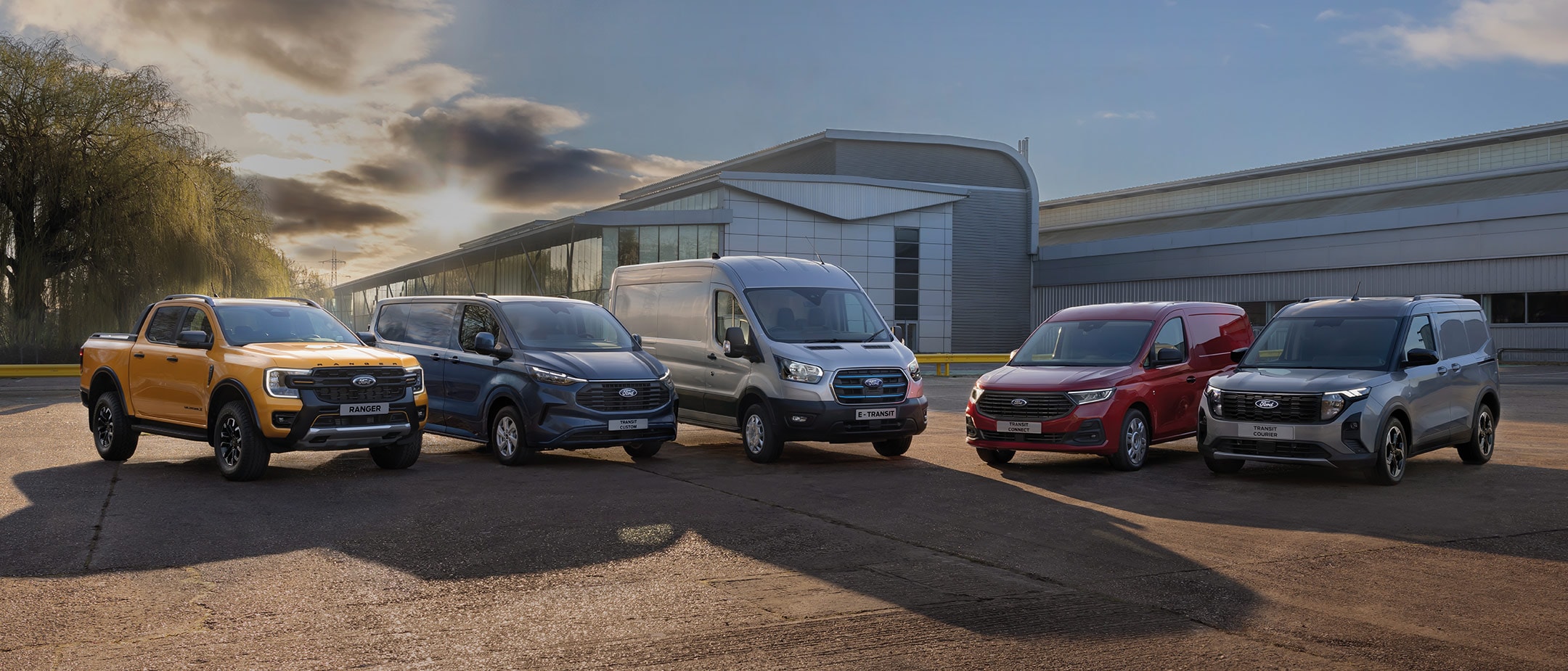 Ford Ranger, Transit Custom, Transit, Transit Connect und Transit Courier parken nebeneinander vor einem Gebäude bei Sonnenuntergang.