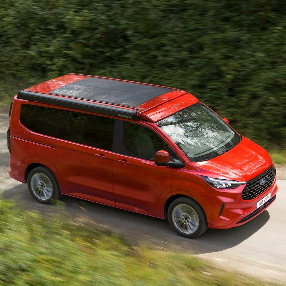 Ford Nugget in Rot. Ansicht von oben, fahrend auf einer Straße. Wald im Hintergrund.