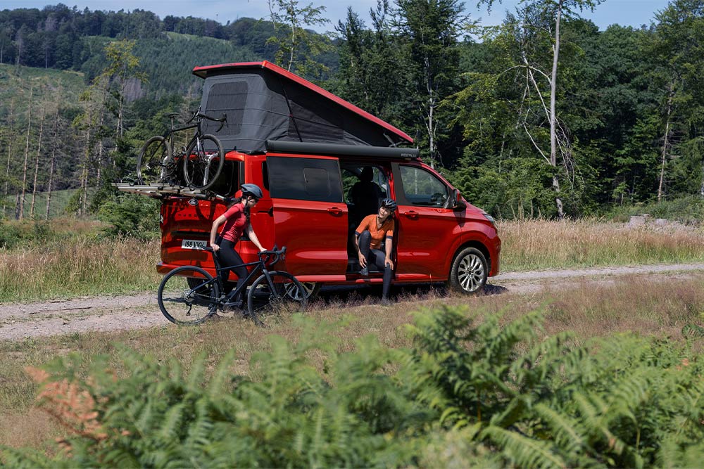 Ford Nugget in Rot. Dreiviertelansicht mit Aufstelldach stehend vor einem Wald. Zwei Personen neben dem Fahrzeug.