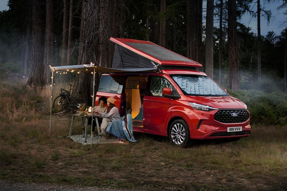 Ford Nugget in Rot. Dreiviertelansicht mit Aufstelldach, parkt in einem Wald.