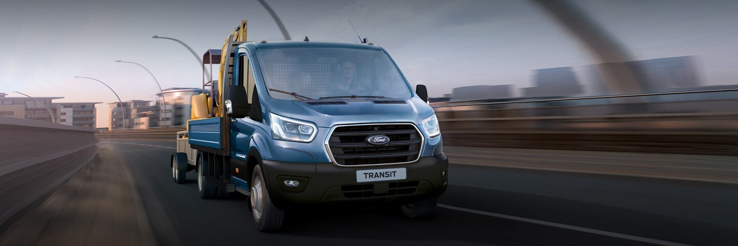 Ford Transit Pritschenwagen mit Anhänger in Blau in der Dreiviertel-Frontansicht auf Straße fahrend.
