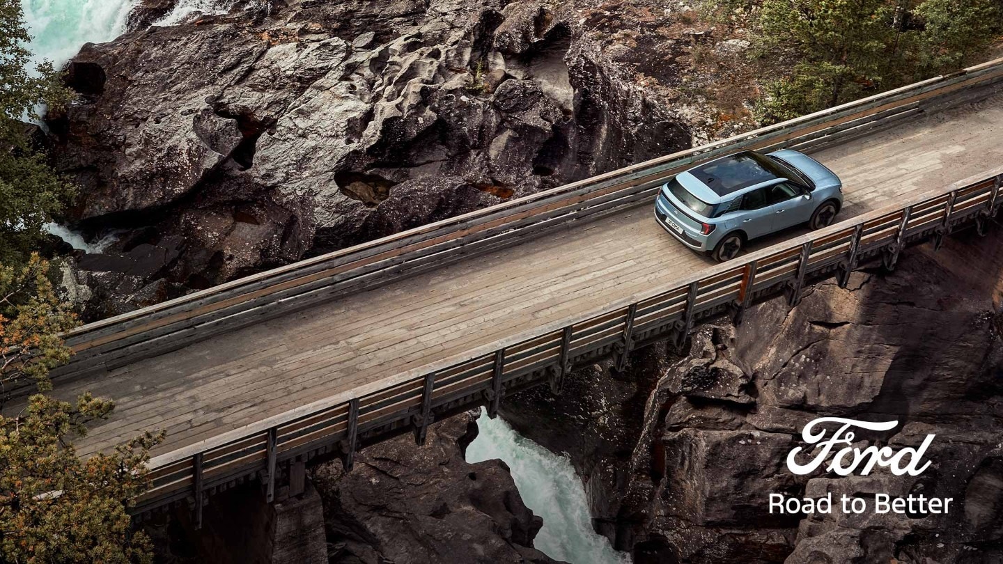 Vollelektrischer Ford Explorer in Blau. Vogelperspektive, fährt über eine schmale Holzbrücke über einen Fluss.