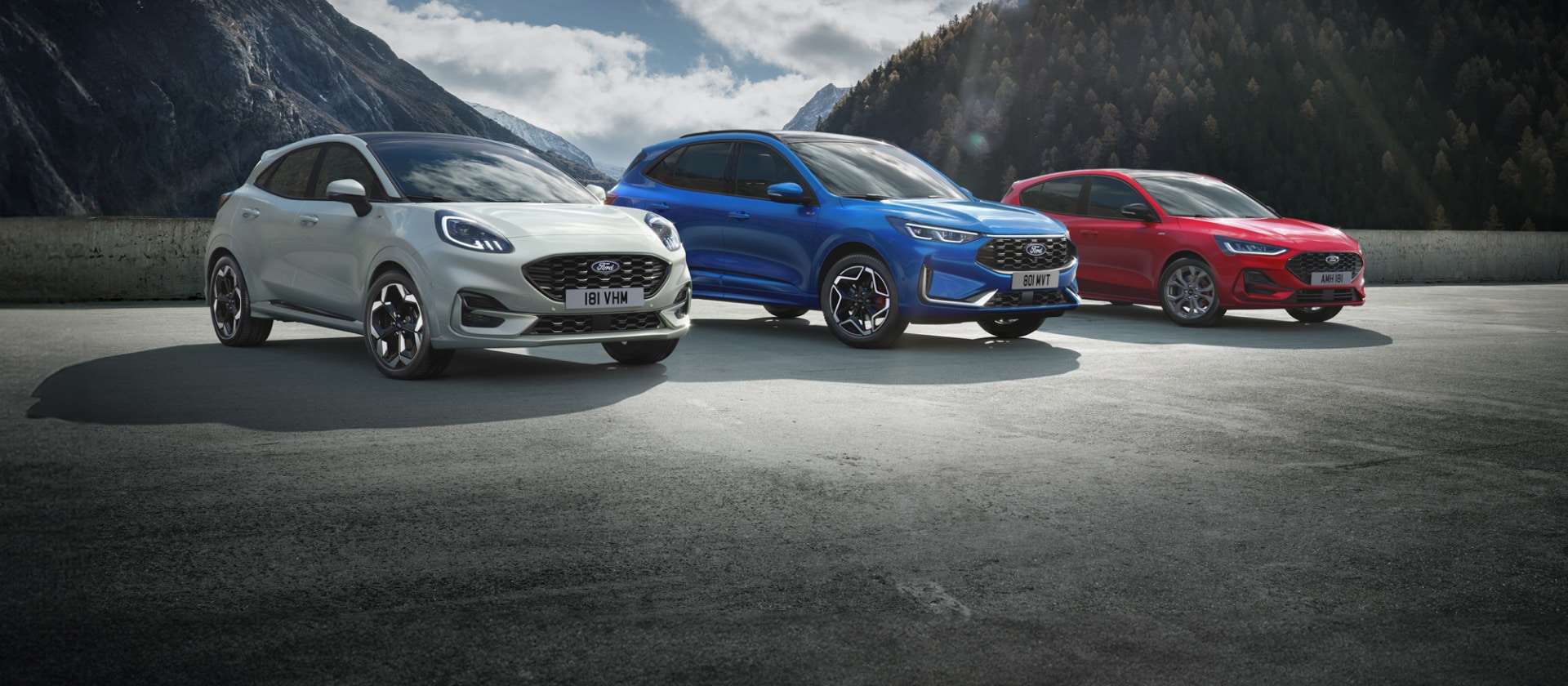 Ford Puma, Ford Kuga und Ford Focus stehen nebeneinander auf einem Parkplatz mit Berglandschaft im Hintergrund.
