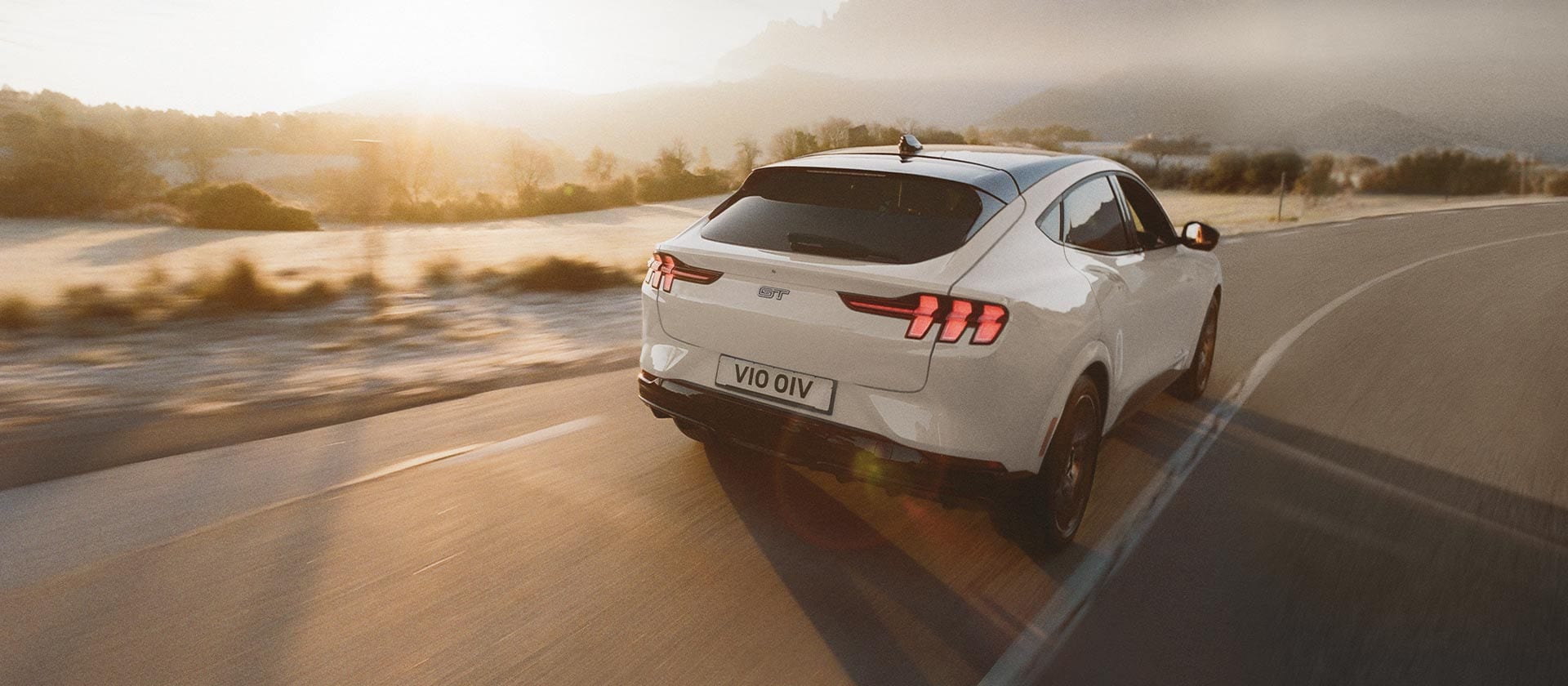 Ford Mustang Mach-E GT in Weiß. Heckansicht, fahrend auf einer Straße bei Sonnenuntergang. Hügel im Hintergrund.