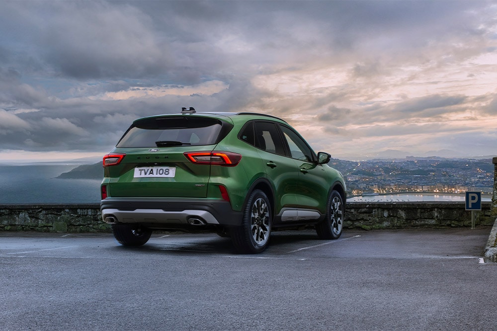 Ford Kuga in Grün. Heckansicht, parkend vor einem Gelände bei Abenddämmerung. 