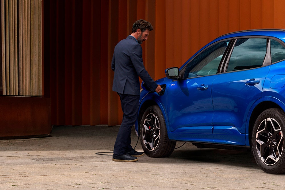Ford Kuga in Blau. Heckansicht, Mann schließt Ladekabel an das Fahrzeug an.