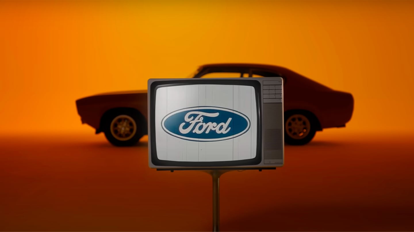 Ford Capri in Gelb. Seitenansicht, stehend vor einer gelben Wand. Fernseher mit Ford-Logo im Vordergrund.