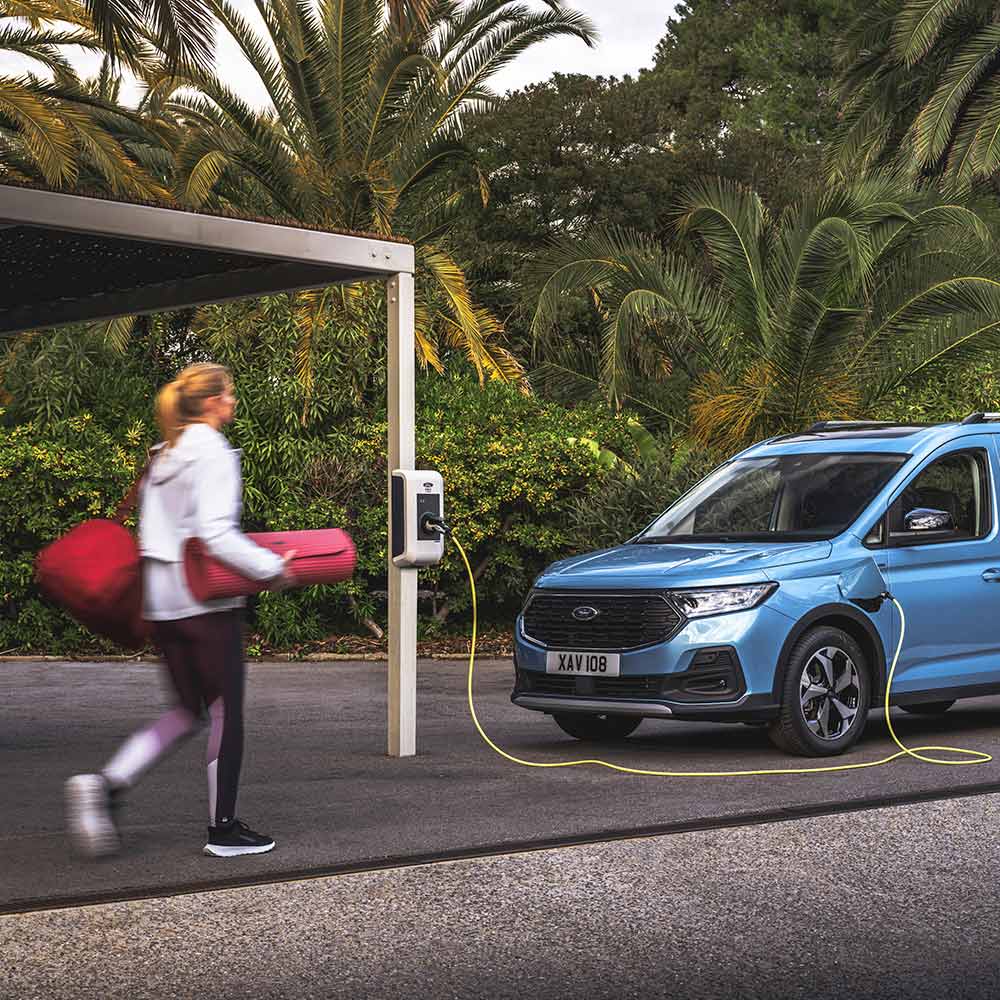 Ford Tourneo Connect in Blau. Dreiviertelansicht, an einer Ladestation angeschlossen. Frau geht zum Fahrzeug.