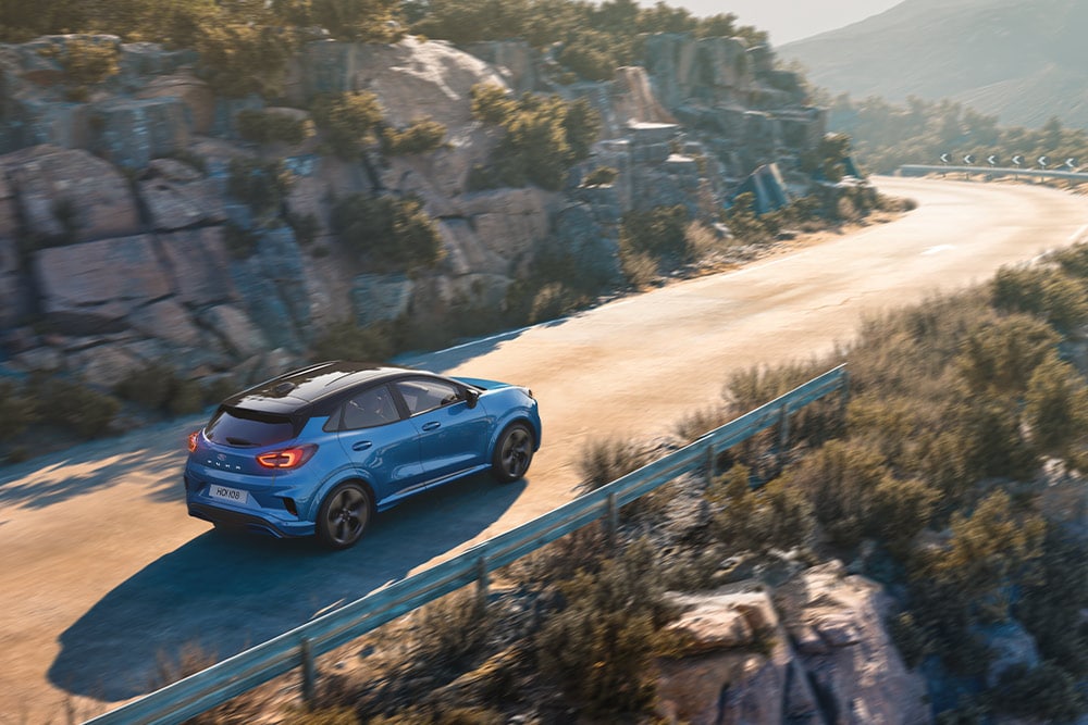 Ford Puma Gen-E in Blau. Dreiviertelansicht, fährt auf einer kurvigen Bergstraße.