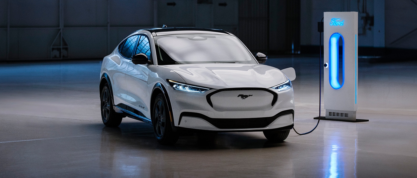 Ford Mustang Mach-E in Weiß. Dreiviertelansicht, ist an einer Ford Ladesäule angeschlossen und lädt.