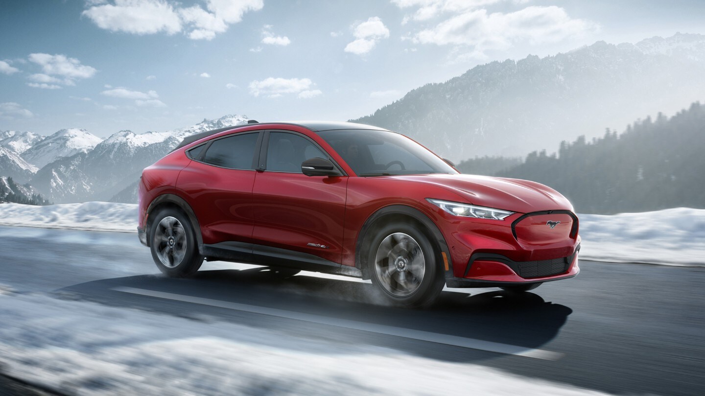 Ford Mustang Mach-E in Rot. Dreiviertelansicht, auf einer Landstraße fahrend mit verschneiter Landschaft