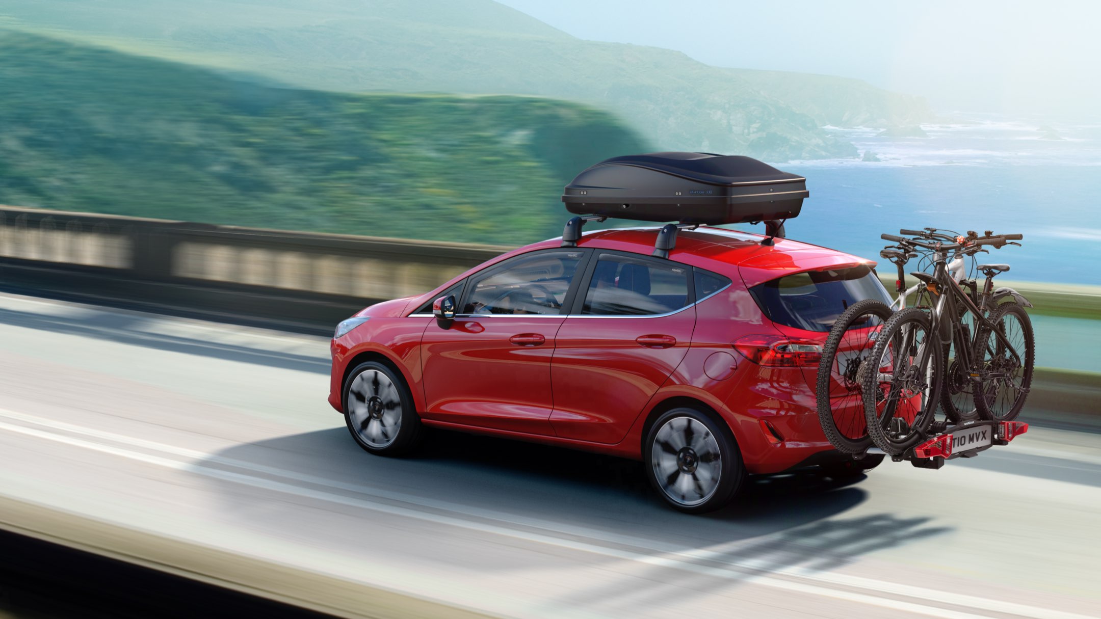 Ford Fiesta in Rot. Dreiviertelansicht, fährt auf einer Brücke mit Dachbox und Heckfahrradträger