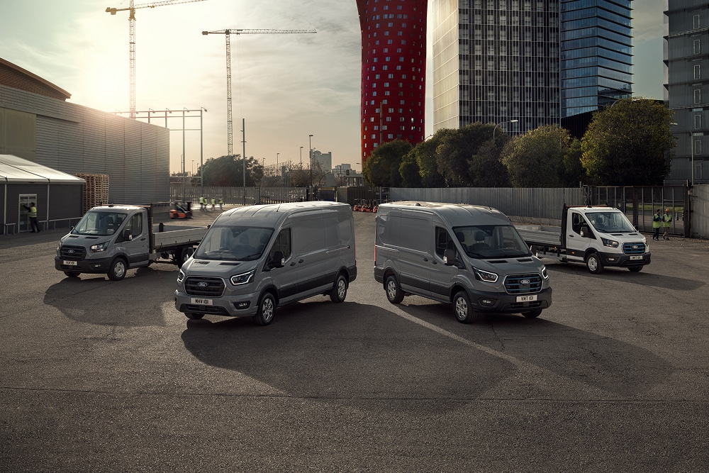 Ford Elektro- und Plug-in-Hybrid-Nutzfahrzeuge auf einem Parkplatz bei Sonnenuntergang, mit Hochhäusern im Hintergrund.