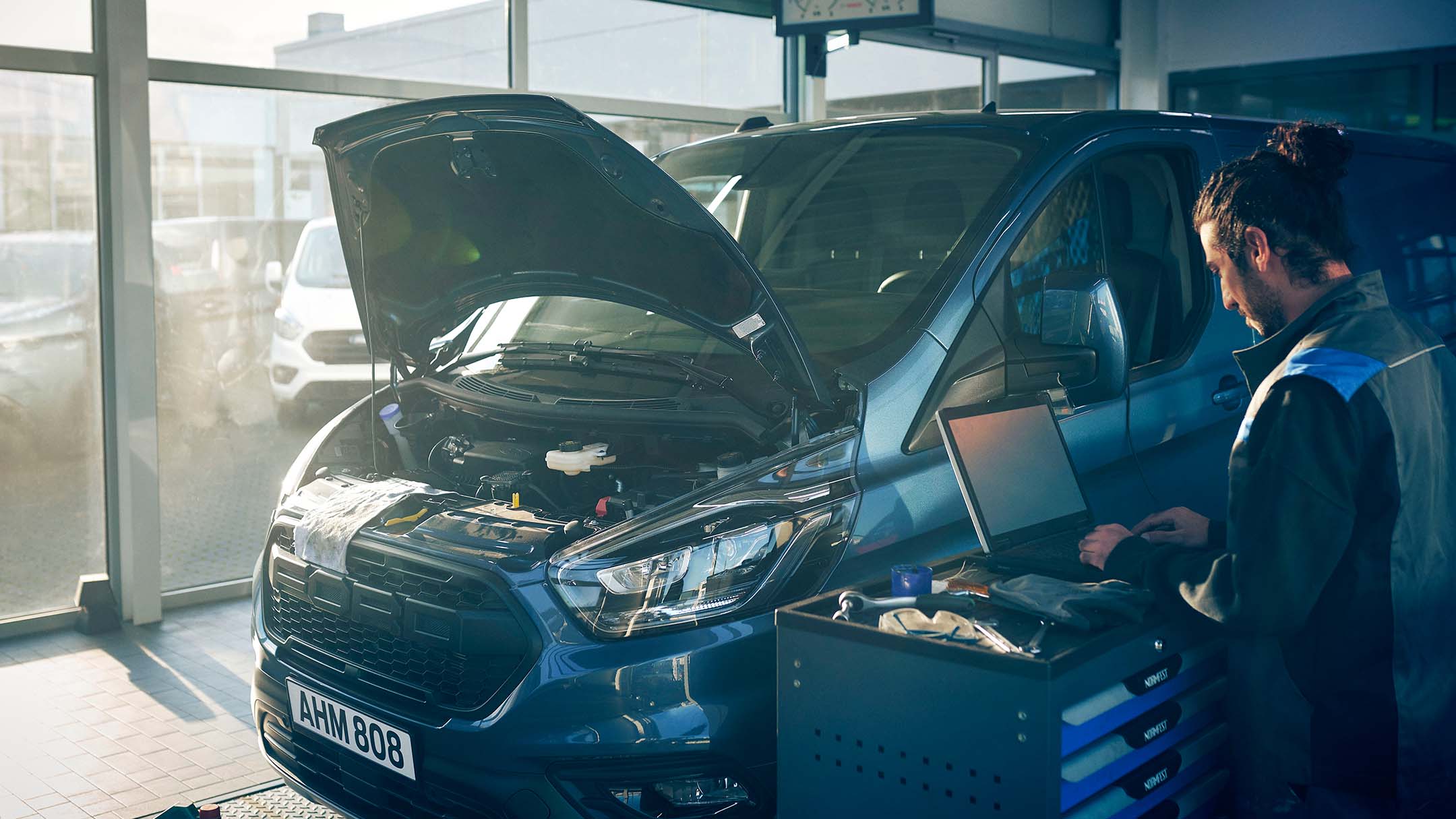 Ein blaues Ford Fahrzeug mit geöffneter Motorhaube steht in einer Werkstatt. Daneben arbeitet eine Person an einem Laptop.
