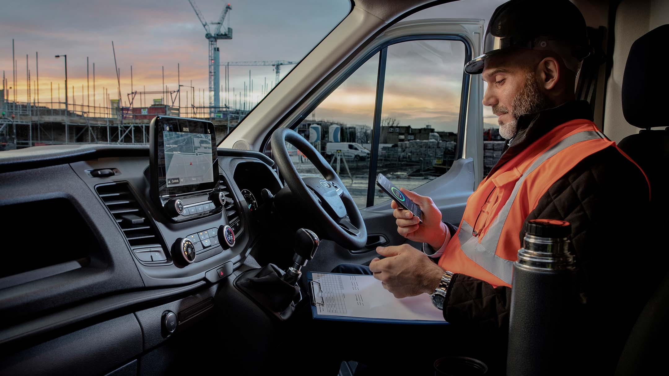 FordliiveArbeiter mit Bauhelm sitzt hinter dem Steuer eines Fahrzeugs und schaut auf ein Mobiltelefon