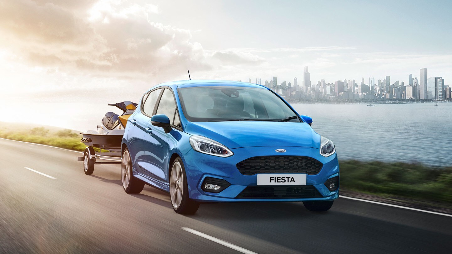 Ford Fiesta in Blau. Frontansicht, fahrend auf Straße mit Anhänger