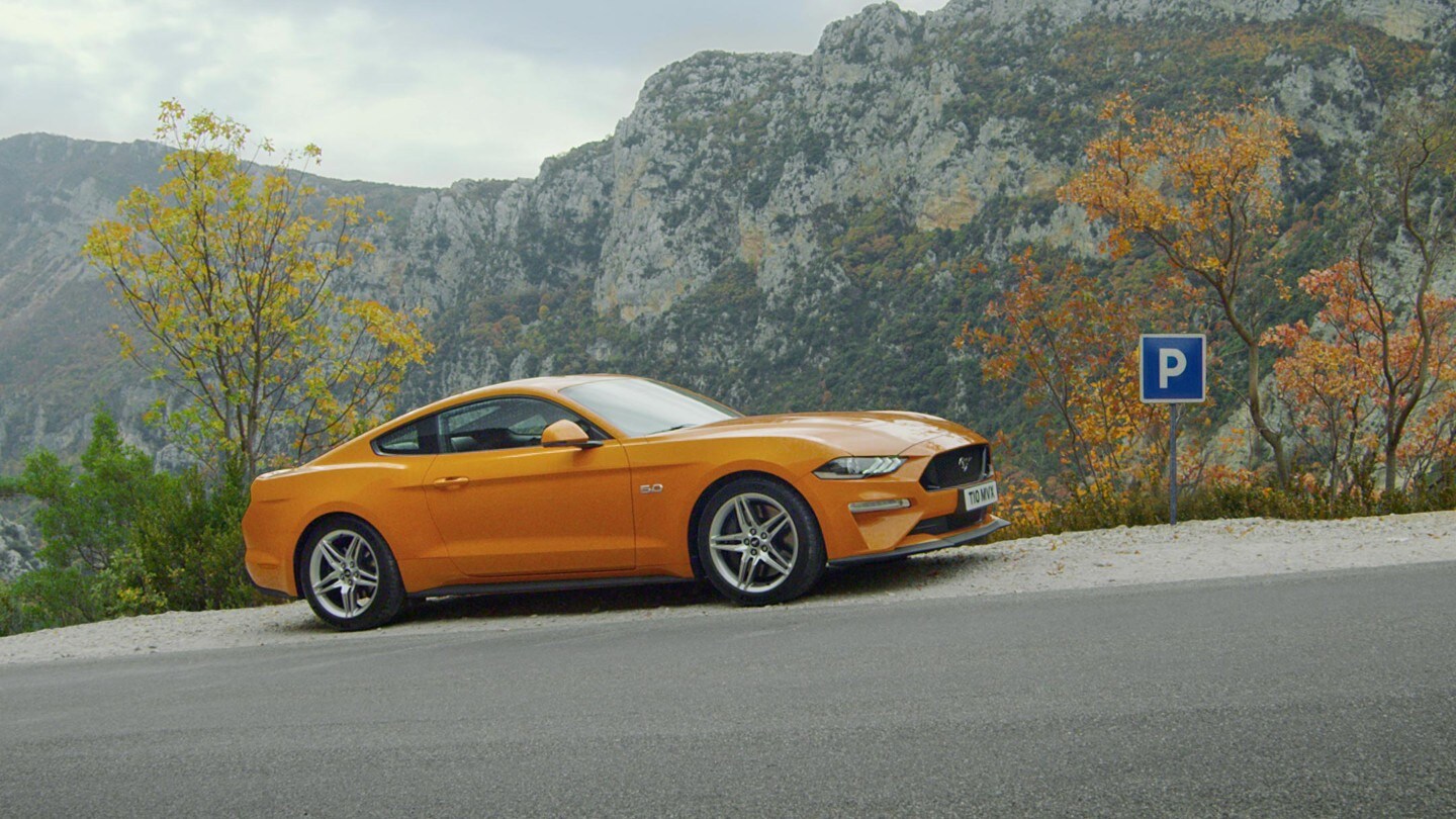 Ford Mustang in Orange. Seitenansicht, parkend auf einer Straße mit Steigung vor einem Gebirge