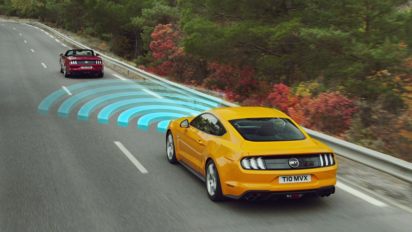 Ford Mustang in Orange. Dreiviertelansicht, fahrend auf einer Straße hinter einem roten Auto mit visualisierter Geschwindigkeitsregelanlage
